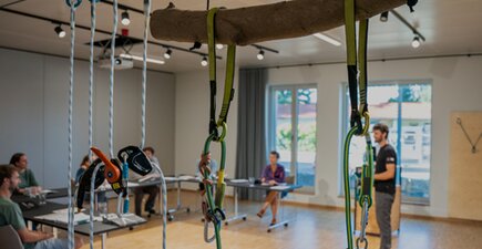 Schulungsraum, verschwommen im Hintergrund sitzen die Schulungsteilnehmer am Tisch, vorne steht ein Schulungsleiter und im Fokus sind einige Arbeitssicherheitsgeräte hängend im Seil zu sehen.